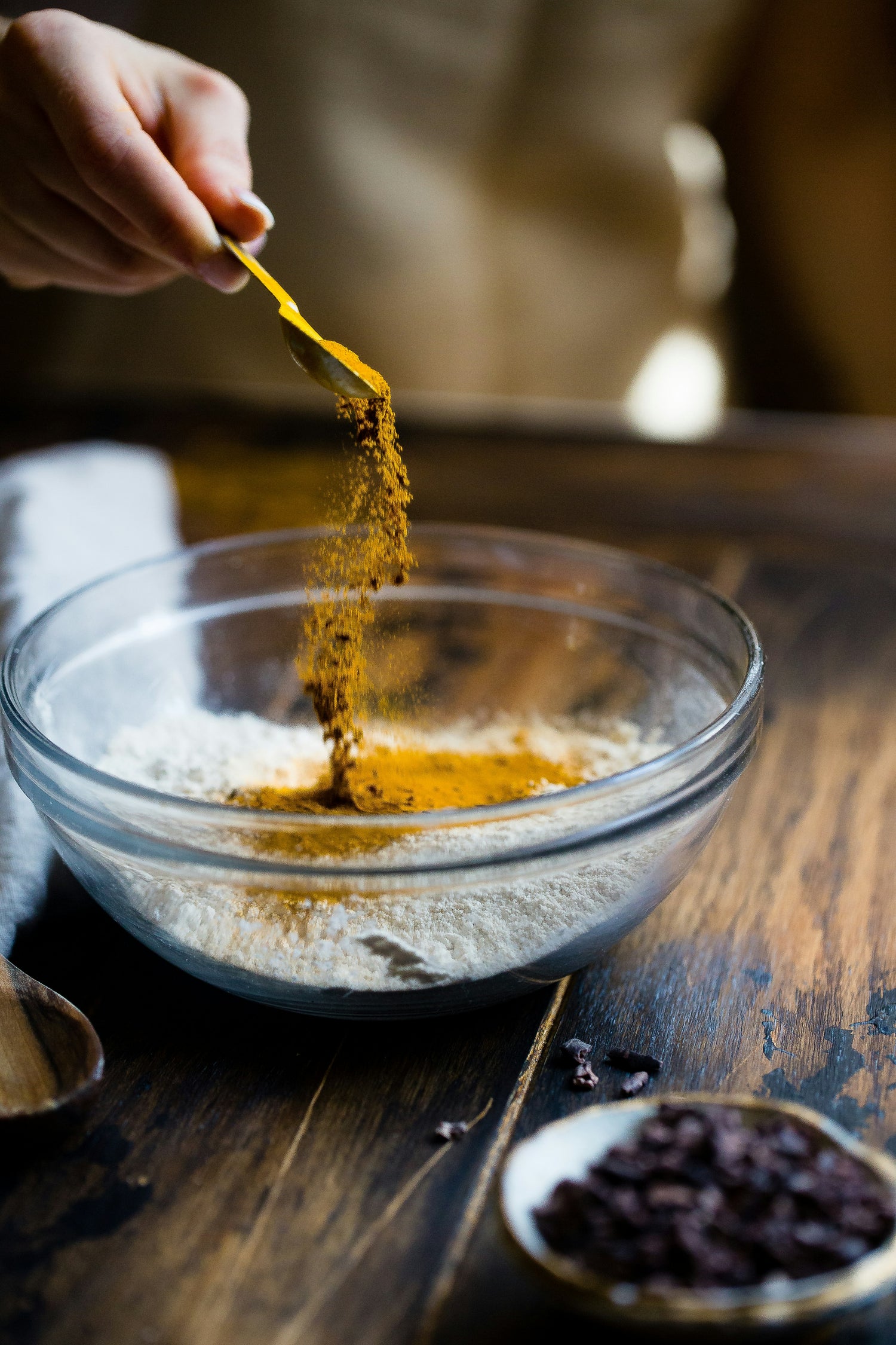 Gemahlener Kurkuma fällt vom Holzlöffel in eine Schale - warm-goldene Gewürzpulveraufnahme.
